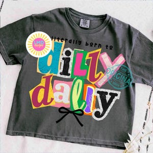 May include: Dark gray t-shirt with the phrase "literally born to dilly dally" in colorful, cut-out style letters. A black bow is at the bottom. The shirt has a vintage, worn look.