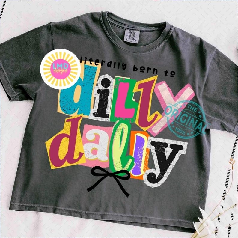 May include: Dark gray t-shirt with the phrase "literally born to dilly dally" in colorful, cut-out style letters. A black bow is at the bottom. The shirt has a vintage, worn look.