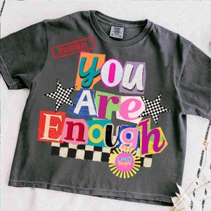 Pu&ograve; includere: T-shirt grigio scuro con la scritta "You Are Enough" in un design colorato in stile collage. Il design include stelle, un motivo a scacchiera e la parola "ORIGINAL" stampata in rosso. La maglietta ha anche un logo.