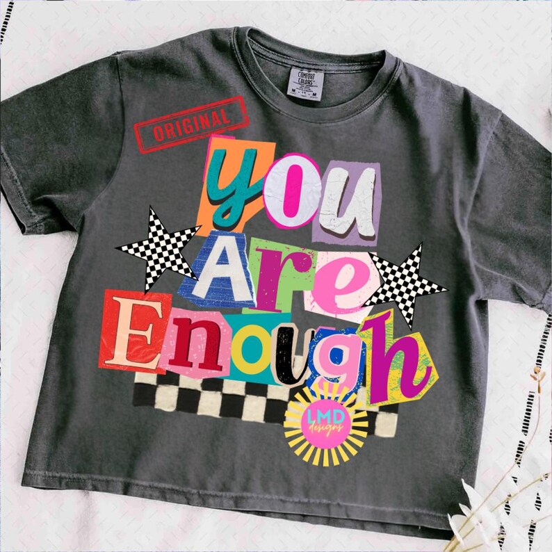 Pu&ograve; includere: T-shirt grigio scuro con la scritta "You Are Enough" in un design colorato in stile collage. Il design include stelle, un motivo a scacchiera e la parola "ORIGINAL" stampata in rosso. La maglietta ha anche un logo.