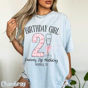 Może przedstawiać: Jasnoniebieska koszulka z napisem "Birthday Girl" i r&oacute;żową cyfrą 2 z kokardkami. Pod cyfrą znajduje się napis "Lauren's 21st Birthday Nashville, TN".