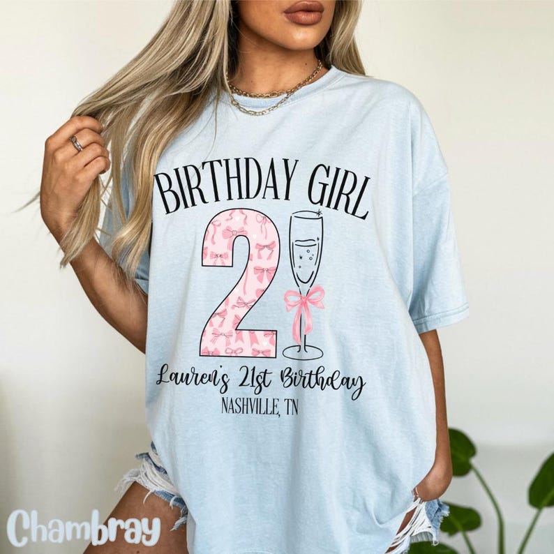 Może przedstawiać: Jasnoniebieska koszulka z napisem "Birthday Girl" i r&oacute;żową cyfrą 2 z kokardkami. Pod cyfrą znajduje się napis "Lauren's 21st Birthday Nashville, TN".