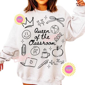 May include: A light gray sweatshirt with a black graphic design. The design includes a crown, push pins, a bow, a globe, a smiley face, a paperclip, an A+ grade, a coffee mug, an apple, a laptop, a pen, and math symbols. The text reads "Queen of the Classroom."