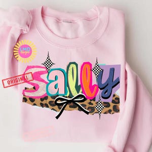 May include: A light pink sweatshirt with the name "Sally" in a colorful, layered design. The letters have a mix of colors, including pink, teal, orange, and blue, with a leopard print and a black bow. The word "ORIGINAL" is printed on a red stamp.