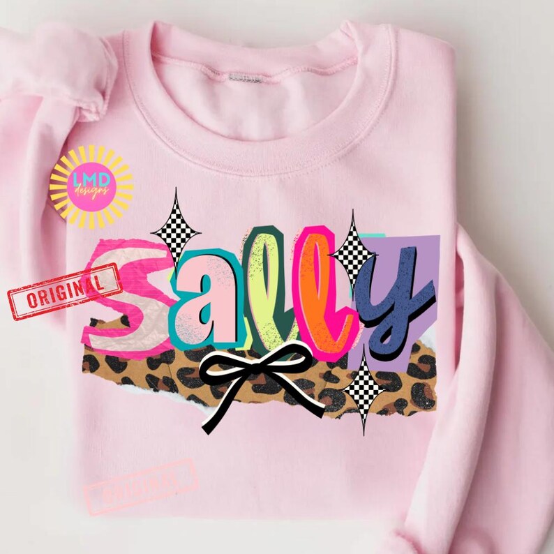 May include: A light pink sweatshirt with the name "Sally" in a colorful, layered design. The letters have a mix of colors, including pink, teal, orange, and blue, with a leopard print and a black bow. The word "ORIGINAL" is printed on a red stamp.