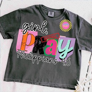 May include: Dark gray t-shirt with the words "girl. PRAY Philippians 4:6" in a colorful, layered design. The word "girl" is in a cursive font, and the word "PRAY" is in a block letter style. A logo is on the upper right.