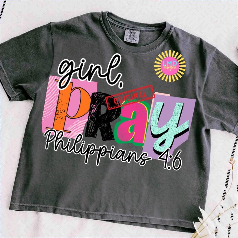 May include: Dark gray t-shirt with the words "girl. PRAY Philippians 4:6" in a colorful, layered design. The word "girl" is in a cursive font, and the word "PRAY" is in a block letter style. A logo is on the upper right.