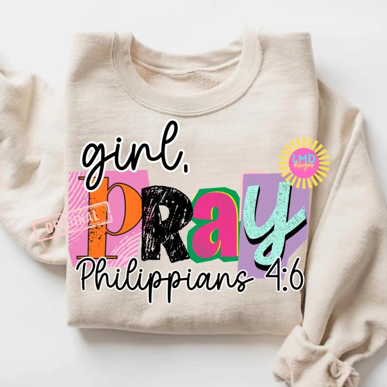 May include: A beige sweatshirt with the words "girl. PRAY Philippians 4:6" in a colorful, block letter design. The word "PRAY" is in black, pink, orange, and teal. The design includes a sun-shaped logo.