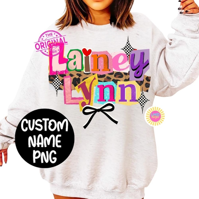 May include: A light gray sweatshirt with a colorful graphic design featuring the names "Lainey Lynn" in various fonts and colors. The design includes a leopard print section, a black bow, and the text "CUSTOM NAME PNG" in a black circle.
