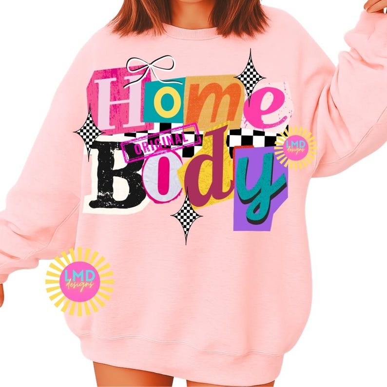 May include: A light pink sweatshirt with the words "Home Body" in a colorful, retro design. The text is layered over various shapes and colors, including pink, teal, purple, and black and white checkered accents. The sweatshirt has long sleeves.