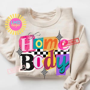 May include: A beige sweatshirt with the words "Home Body" in a colorful, layered design. The design includes pink, orange, blue, and purple letters, with a black and white checkered pattern and a bow. The word "ORIGINAL" is stamped on the design.