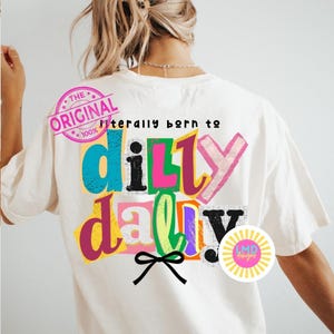 May include: Cream-colored t-shirt with the text "Literally born to dilly dally" in colorful, layered letters. The design includes a black bow and a pink stamp that says "The Original". A circular logo with the text "LMD designs" is also visible.