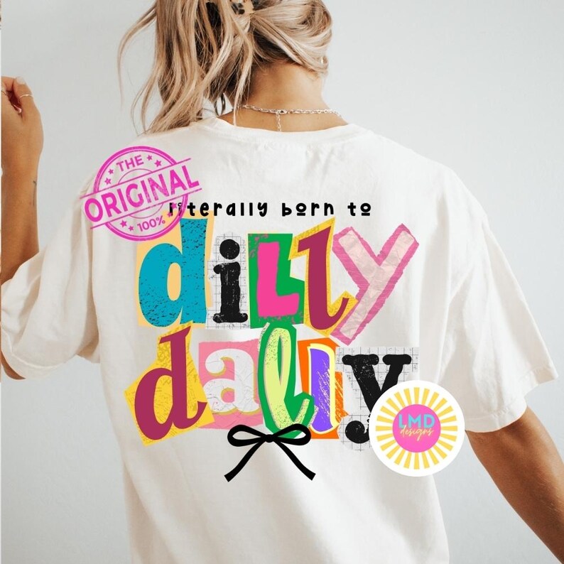 May include: Cream-colored t-shirt with the text "Literally born to dilly dally" in colorful, layered letters. The design includes a black bow and a pink stamp that says "The Original". A circular logo with the text "LMD designs" is also visible.