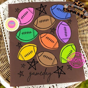May include: A brown t-shirt featuring a colorful graphic print of multiple footballs in pink, blue, brown, green, orange, purple, yellow, and lime green. The design includes black hand-drawn stars and the word gameday in cursive script.