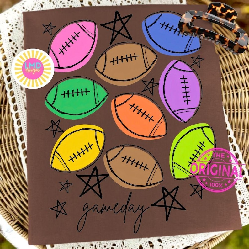May include: A brown t-shirt featuring a colorful graphic print of multiple footballs in pink, blue, brown, green, orange, purple, yellow, and lime green. The design includes black hand-drawn stars and the word gameday in cursive script.