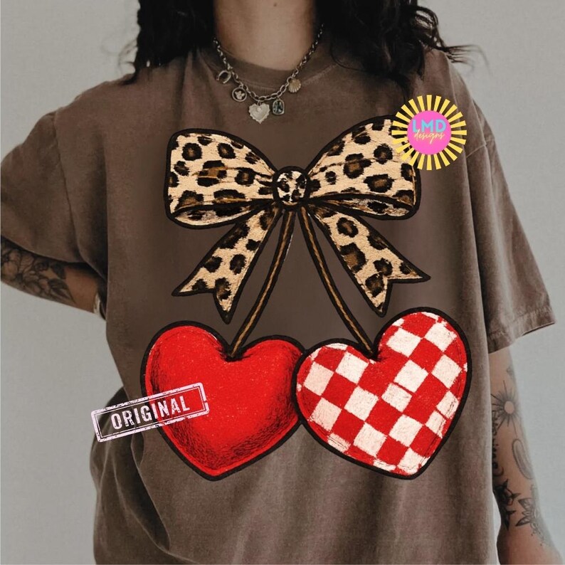May include: Brown t-shirt featuring a leopard print bow above two heart-shaped cherries. One heart is solid red, and the other has a red and white checkered pattern. The word "ORIGINAL" is printed on a small rectangle.