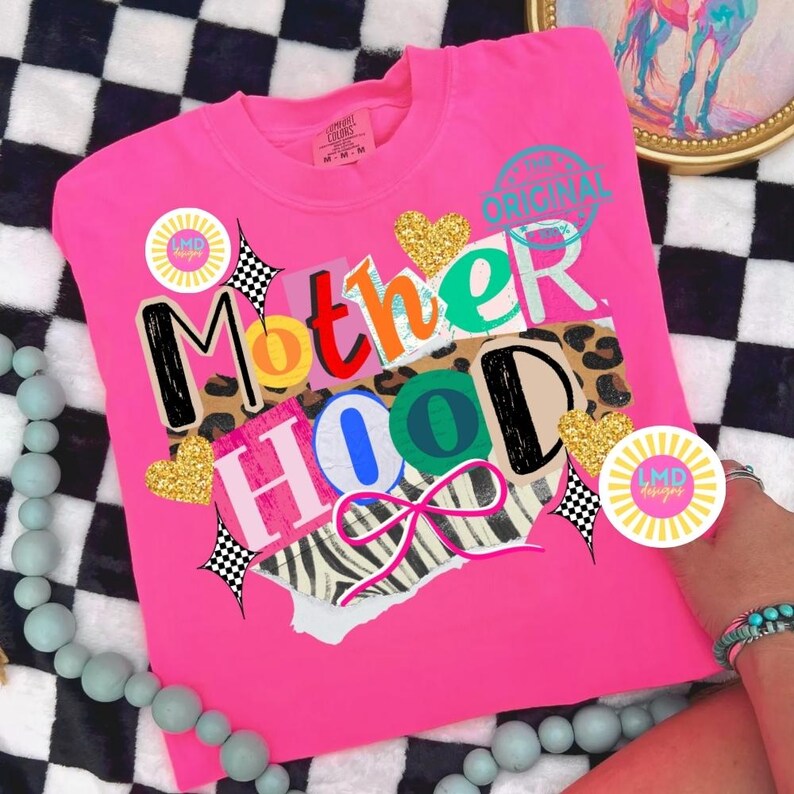 May include: Bright pink t-shirt with the word "Motherhood" in a collage style design. The design includes leopard print, zebra print, gold glitter hearts, and a pink bow. The shirt has a crew neck and short sleeves.