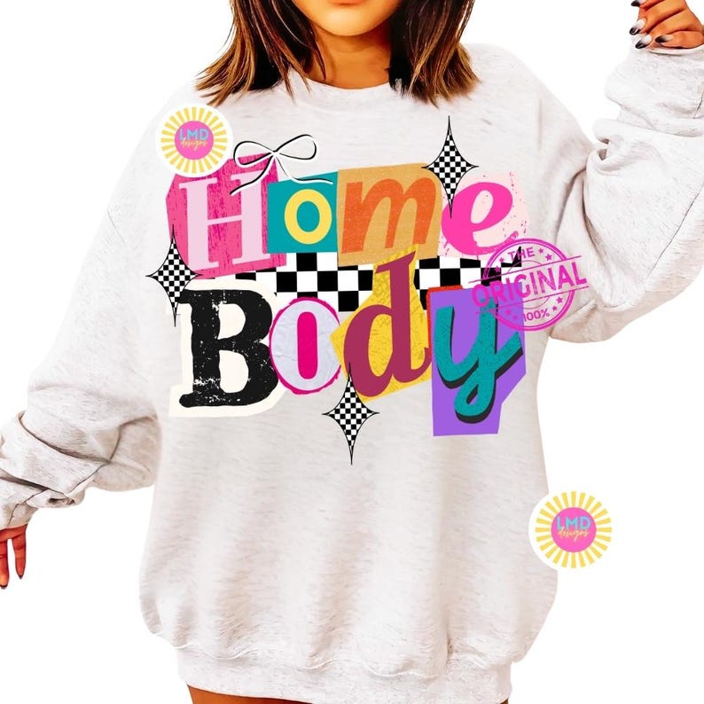 May include: A light gray sweatshirt with the words "Home Body" in a colorful, layered design. The text includes pink, orange, teal, and purple hues, with black and white checkered accents. The design also features a bow and starbursts.