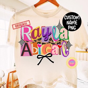 May include: Cream-colored t-shirt with the name "Rayla Abigail" in colorful, layered text. The design includes a leopard print background, hearts, and a black bow. The word "ORIGINAL" is stamped in red.