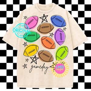 May include: A beige short-sleeve t-shirt featuring a graphic print of colorful footballs in pink, blue, green, orange, yellow, and brown, interspersed with black star outlines and the handwritten text "gameday" in black script.