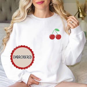 May include: White sweatshirt with a red cherry design embroidered on the front. The sweatshirt has a round, red label with the word "EMBROIDERED" in white text.