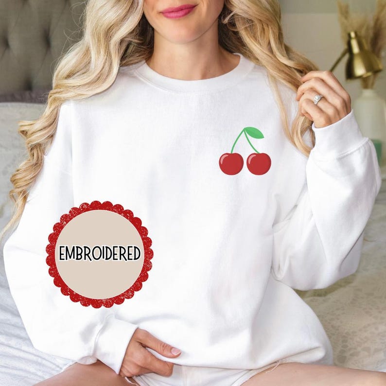 May include: White sweatshirt with a red cherry design embroidered on the front. The sweatshirt has a round, red label with the word "EMBROIDERED" in white text.
