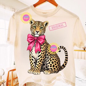 May include: Cream-colored t-shirt featuring a seated leopard with a large pink bow. The leopard has a spotted coat and green eyes. The shirt also has the word "ORIGINAL" and the text "LMD designs" on it.