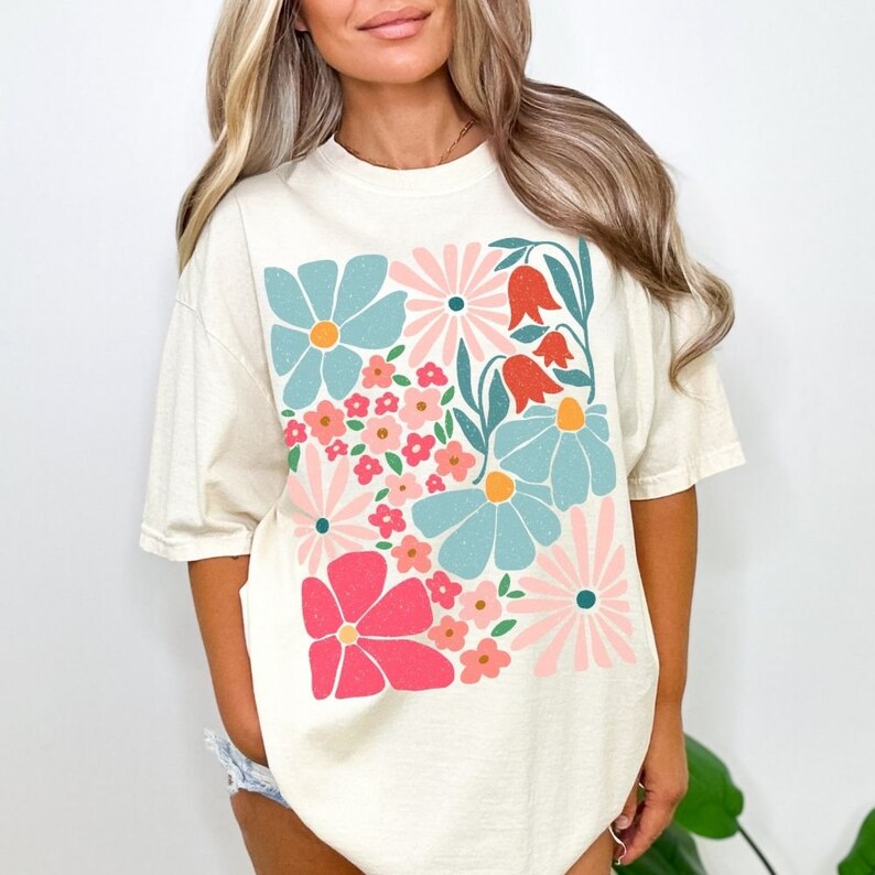 K&ouml;nnte beinhalten: Ein cremefarbenes T-Shirt mit einem bunten Blumenmuster. Der Druck zeigt rosa, blaue, rote und gr&uuml;ne Blumen.