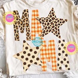 May include: A cream-coloured t-shirt featuring the word MAMA in large letters. The letters are decorated with leopard print, orange and white gingham, and brown polka dots. Two large stars are integrated into the design, and two yellow sunburst logos with the text LMD Designs are visible.