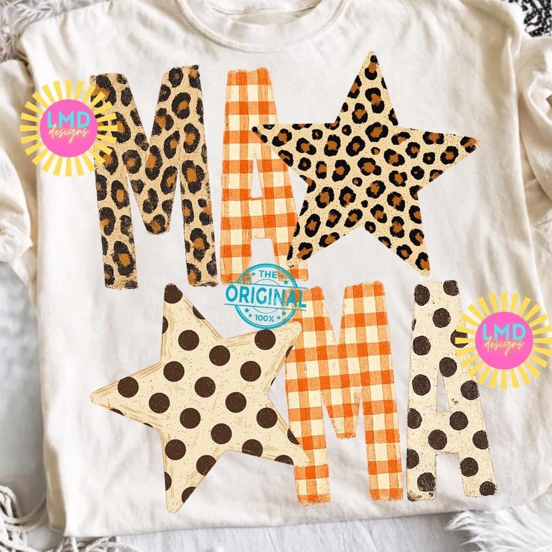 May include: A cream-coloured t-shirt featuring the word MAMA in large letters. The letters are decorated with leopard print, orange and white gingham, and brown polka dots. Two large stars are integrated into the design, and two yellow sunburst logos with the text LMD Designs are visible.