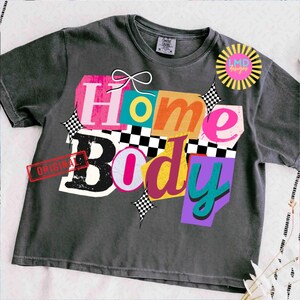 May include: Dark gray t-shirt with the words "Home Body" in a colorful, collage-style design. The design includes pink, orange, yellow, and teal letters, with a white bow and checkered accents. A red "ORIGINAL" stamp is visible.