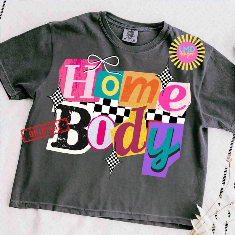 May include: Dark gray t-shirt with the words "Home Body" in a colorful, collage-style design. The design includes pink, orange, yellow, and teal letters, with a white bow and checkered accents. A red "ORIGINAL" stamp is visible.