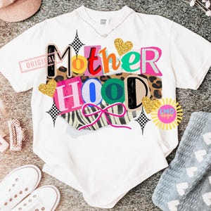 May include: White t-shirt with the word "Motherhood" in a colorful, collage-style design. The design includes leopard print, zebra stripes, and glitter hearts. The shirt has a relaxed fit and a crew neck.