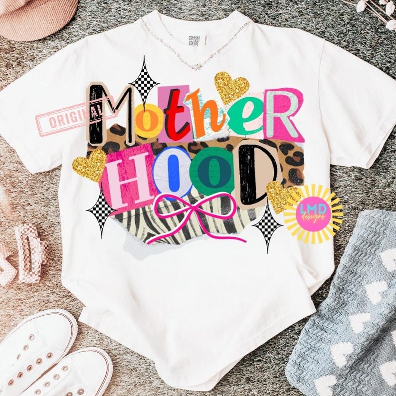 May include: White t-shirt with the word "Motherhood" in a colorful, collage-style design. The design includes leopard print, zebra stripes, and glitter hearts. The shirt has a relaxed fit and a crew neck.