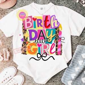 May include: White t-shirt with a colorful "Birthday Girl" graphic. The design features the words "Birthday Girl" in various colors and fonts, with stars, hearts, and a bow. The shirt has a crew neck and short sleeves.