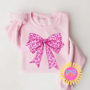 May include: A light pink sweatshirt featuring a large, glittery pink bow with a leopard print design. The bow is centered on the front of the shirt. The sweatshirt has long sleeves and a crew neck.