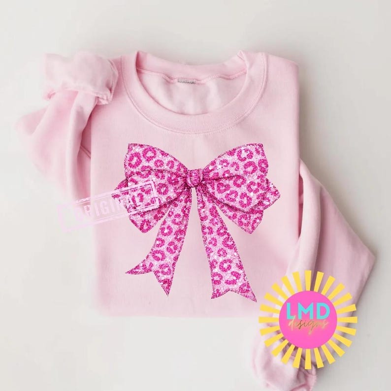 May include: A light pink sweatshirt featuring a large, glittery pink bow with a leopard print design. The bow is centered on the front of the shirt. The sweatshirt has long sleeves and a crew neck.