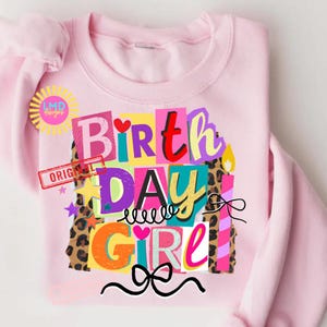 May include: A light pink sweatshirt with a colorful graphic that says "Birthday Girl." The design includes the words "Birth," "Day," and "Girl" in various bright colors and fonts, with leopard print accents and a bow.
