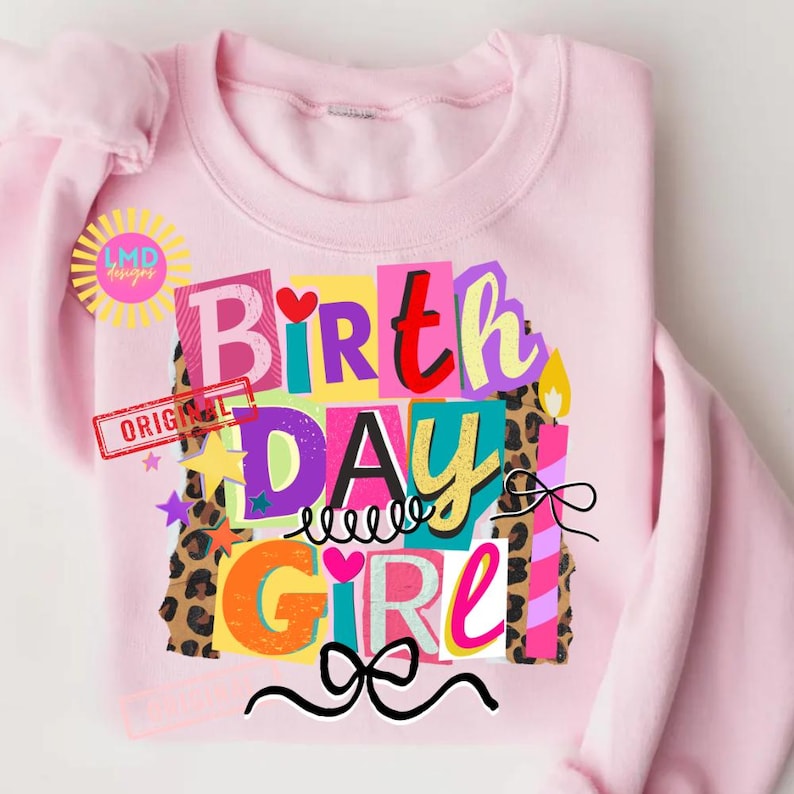 May include: A light pink sweatshirt with a colorful graphic that says "Birthday Girl." The design includes the words "Birth," "Day," and "Girl" in various bright colors and fonts, with leopard print accents and a bow.
