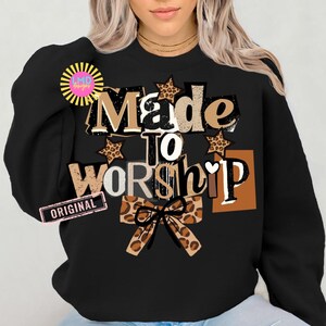 K&ouml;nnte beinhalten: Schwarzes Sweatshirt mit dem Text "Made to Worship" in einer Mischung aus Schriftarten und Farben, einschlie&szlig;lich braunen, wei&szlig;en und Leopardenmuster-Akzenten. Eine Schleife mit Leopardenmuster befindet sich unten. Das Sweatshirt hat einen Rundhalsausschnitt.
