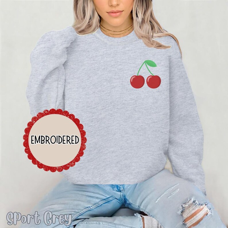 May include: A gray sweatshirt with an embroidered cherry design. The text "EMBROIDERED" is on a red circle label. The sweatshirt is labeled "Sport Grey".