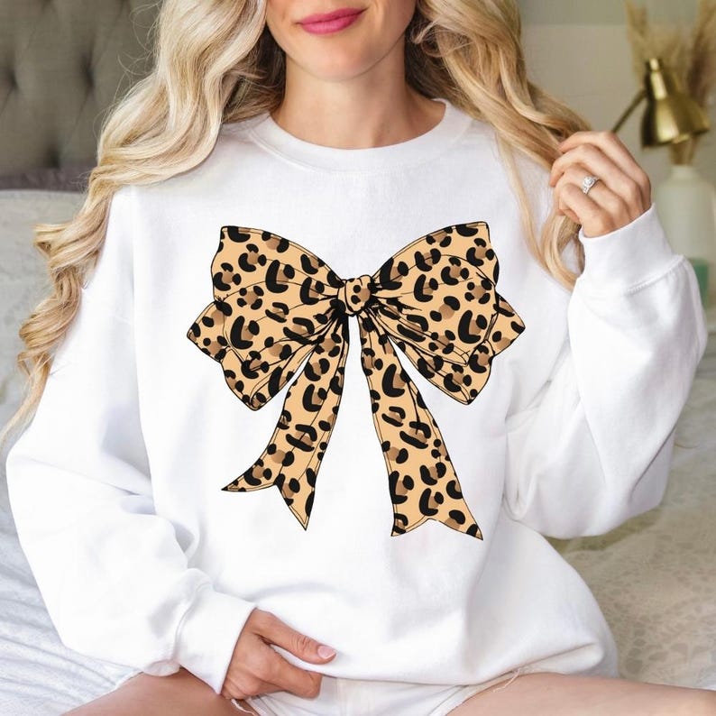 May include: White sweatshirt featuring a large bow design with a leopard print pattern in shades of tan and black. The bow is centered on the front of the long-sleeved top. The sweatshirt is made of a soft material.
