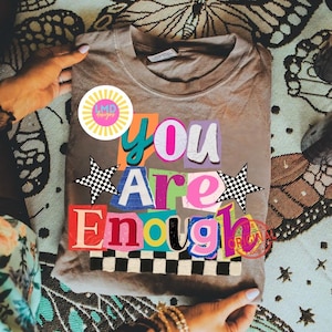 Pu&ograve; includere: Una maglietta color cachi con la scritta "You Are Enough" in un design colorato in stile collage. Il design include stelle, un motivo a scacchiera e un logo circolare. La maglietta &egrave; piegata e tenuta tra le mani.