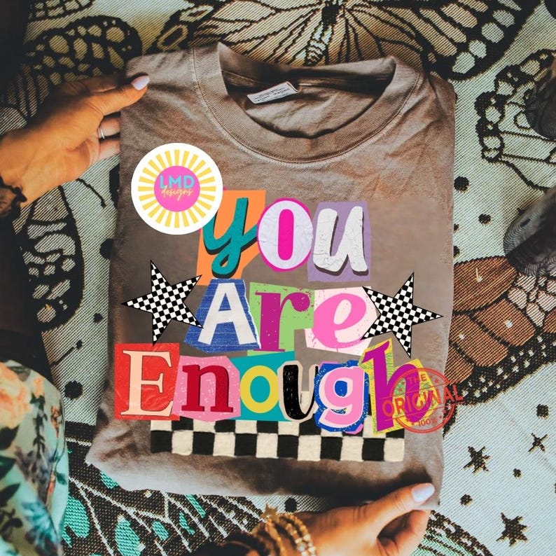 Pu&ograve; includere: Una maglietta color cachi con la scritta "You Are Enough" in un design colorato in stile collage. Il design include stelle, un motivo a scacchiera e un logo circolare. La maglietta &egrave; piegata e tenuta tra le mani.