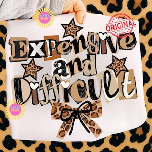 May include: A white t-shirt featuring the text "Expensive and Difficult" in a collage design. The design incorporates leopard print stars and a bow, with a red stamp reading "The Original".