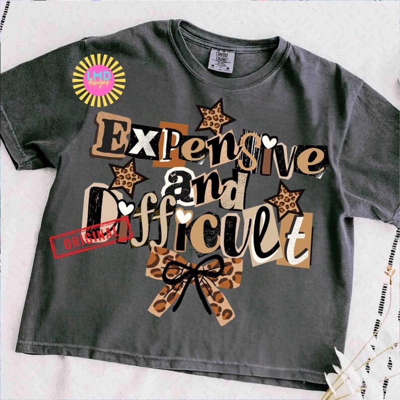 Pu&ograve; includere: T-shirt grigio scuro con la scritta "Expensive and Difficult" in un design a strati. Il design include stelle e cuori, e un fiocco con stampa leopardata. La maglietta ha un girocollo e maniche corte.