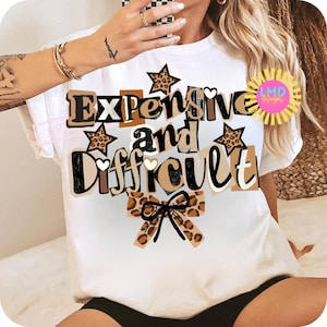 Pu&ograve; includere: T-shirt bianca con la scritta "Expensive and Difficult" in un mix di font e colori, tra cui accenti marroni, neri e stampa leopardata. Il design include stelle e un fiocco.