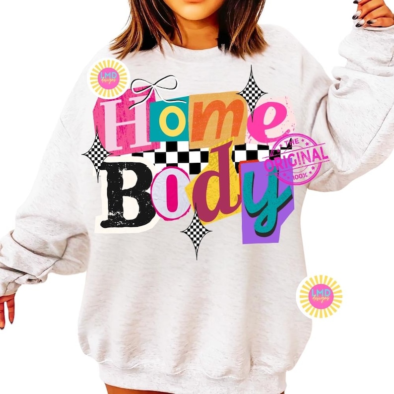 May include: A light gray sweatshirt with the words "Home Body" in a colorful, layered design. The text includes pink, orange, teal, and purple hues, with black and white checkered accents. The design also features a bow and starbursts.