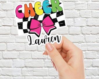 May include: A colorful "CHEER" sticker with a pink bow and a black and white checkered background. The sticker also includes the name "Lauren" in a white cursive font.