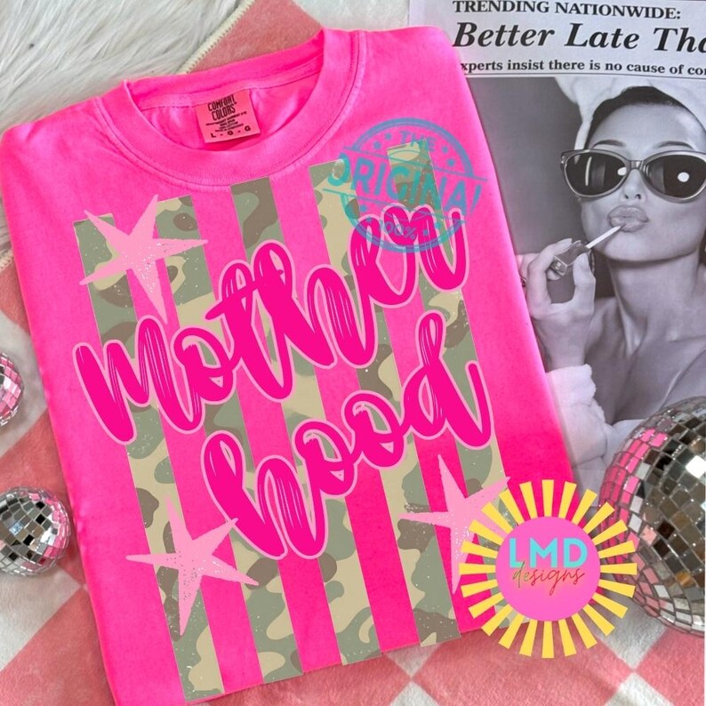 May include: A bright pink sweatshirt with the word "Motherhood" in a cursive font. The design includes vertical stripes, camouflage patterns, and star accents. The text "Original" is also visible, along with the "LMD designs" logo.